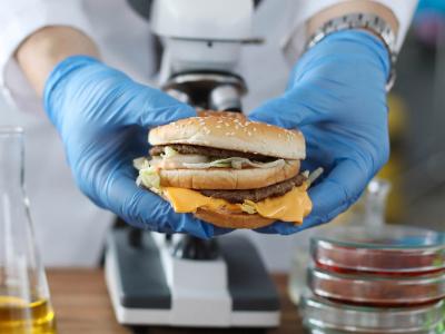 Nahaufnahme von zwei Händen in blauen Handschuhen vor Elektronenmikroskop. Die Hände halten einen Hamburger.